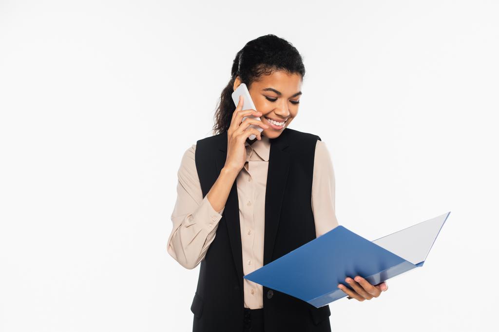 Fröhliche afrikanisch-amerikanische Geschäftsfrau telefoniert und hält Papiermappe isoliert auf weißem Papier  - Foto, Bild