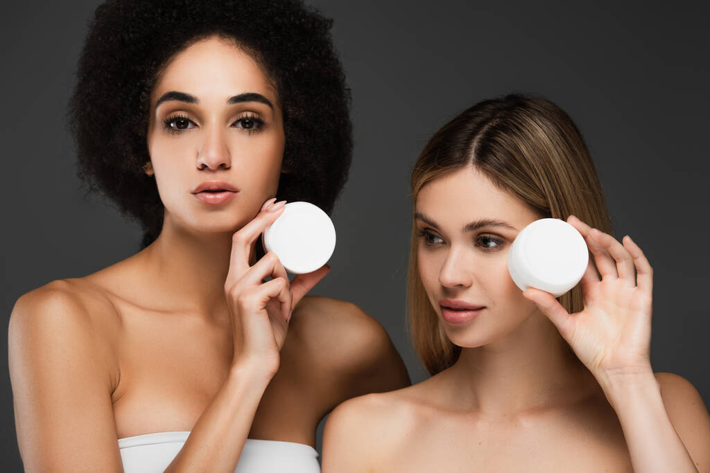 mujeres multiétnicas con piel perfecta mostrando recipientes con crema cosmética aislada en gris - Foto, Imagen