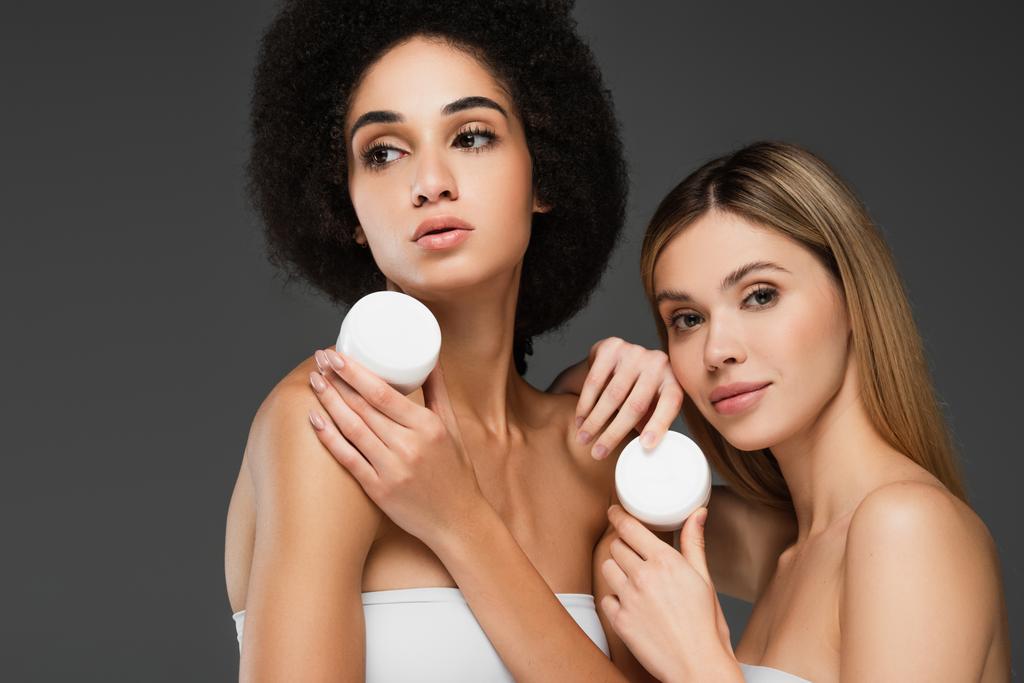 mulheres jovens e muito multiétnicas com pele perfeita e maquiagem natural posando com creme cosmético isolado em cinza - Foto, Imagem