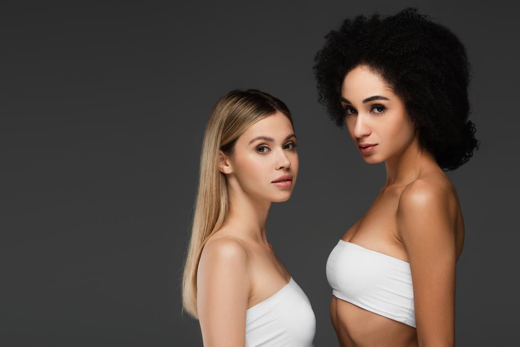 junge multiethnische Frauen in weißen Tops und natürlichem Make-up schauen vereinzelt in die Kamera auf grau - Foto, Bild