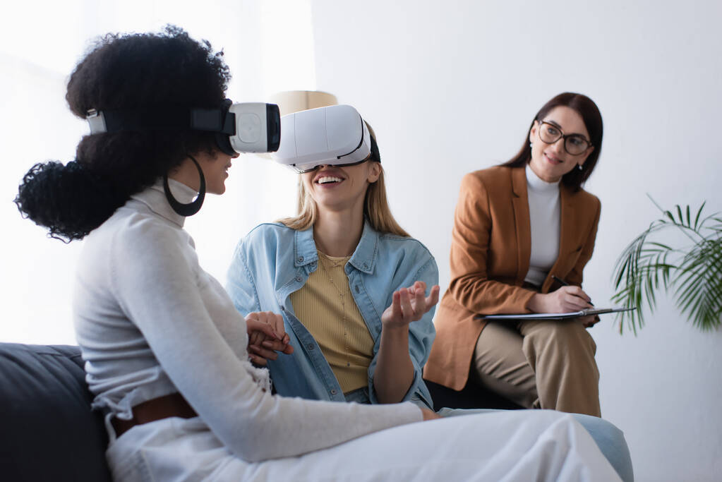 happy interracial lesbian couple in vr headsets holding hands near blurred psychologist writing on clipboard - Photo, Image