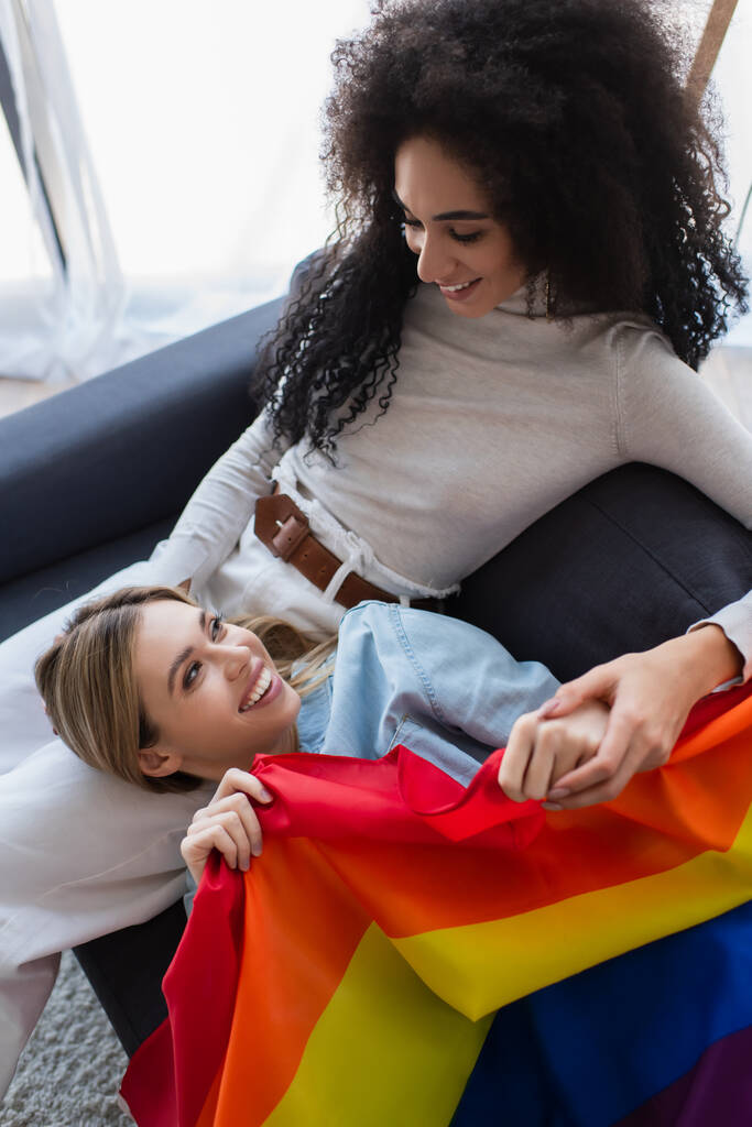 glückliche interrassische lesbische Frauen, die einander auf dem Sofa in der Nähe der lgbt-Flagge anschauen - Foto, Bild