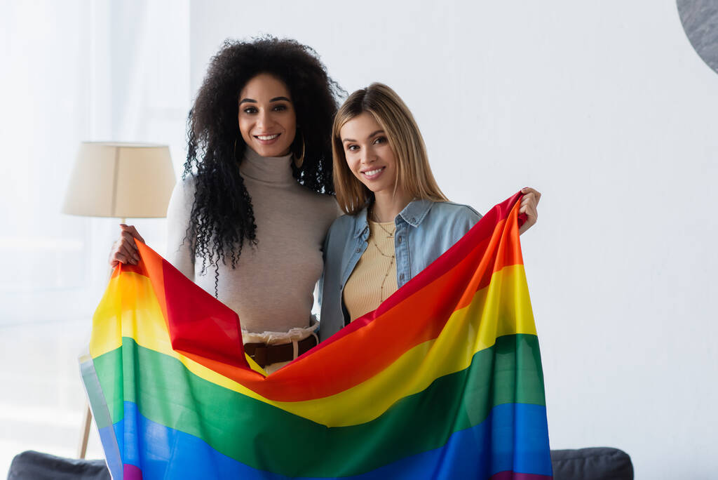 jóvenes y alegres lesbianas multiétnicas mirando a la cámara mientras están de pie con la bandera lgbt - Foto, imagen