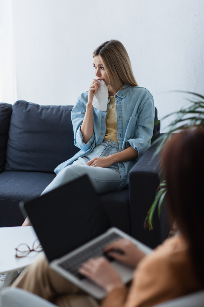 depressed woman sitting on couch near psychologist with laptop on blurred foreground - Photo, Image