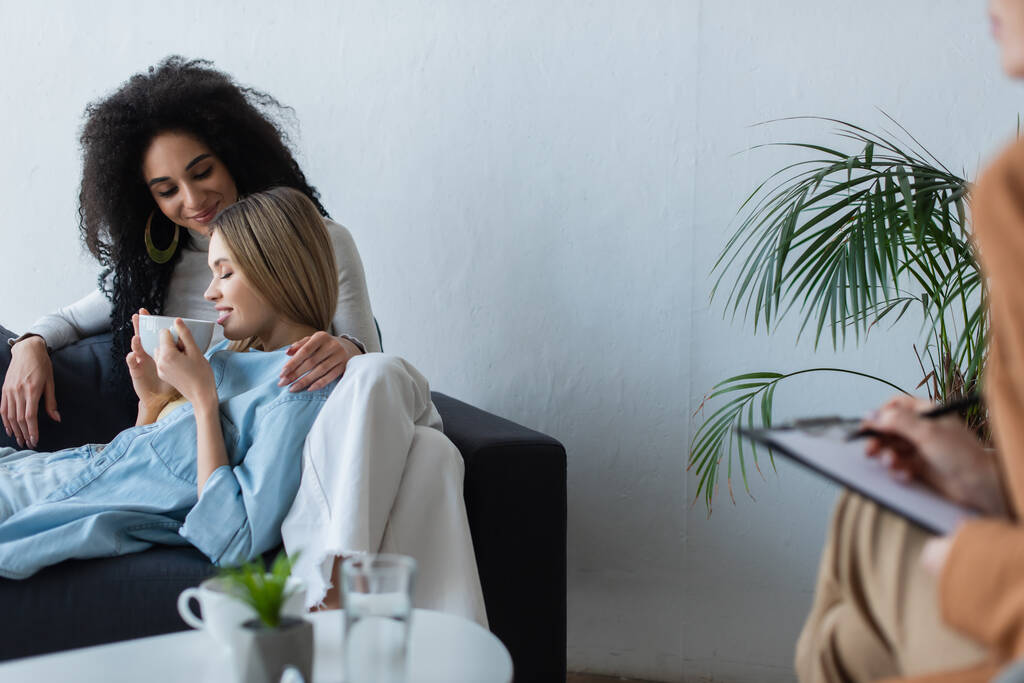 usmívající se lesbičky žena pití čaje na gauči v blízkosti africké americké přítelkyně a rozmazané psycholog psaní na schránce - Fotografie, Obrázek
