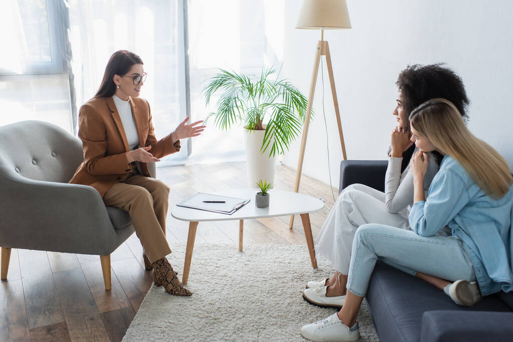 psicólogo positivo gesticulando mientras habla con pareja interracial de lesbianas en la sala de consulta - Foto, imagen