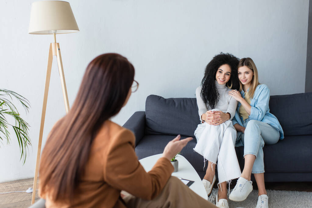 psicóloga borrosa señalando con la mano mientras habla con lesbianas multiétnicas felices sentadas en el sofá - Foto, imagen