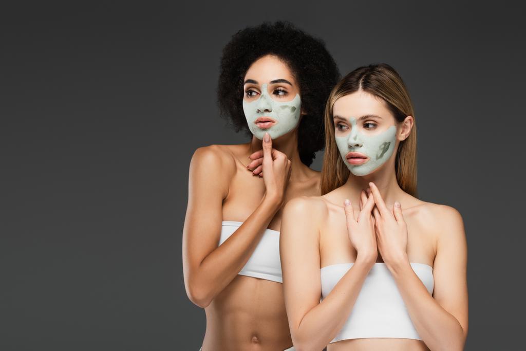 slim interracial women in white tops and clay masks standing isolated on grey - Photo, Image