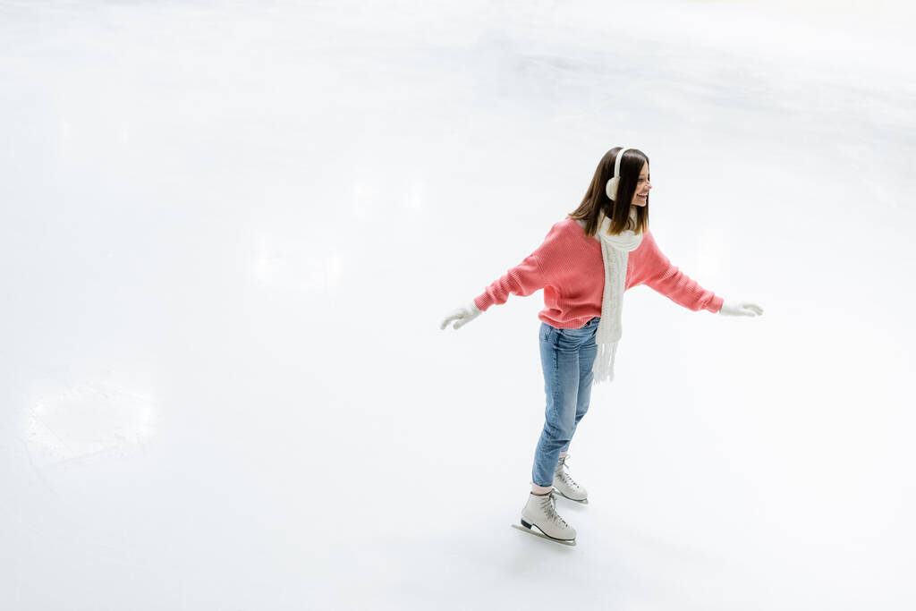 hoge hoek uitzicht van vrolijke jonge vrouw in oor muffs en sjaal schaatsen op de ijsbaan - Foto, afbeelding