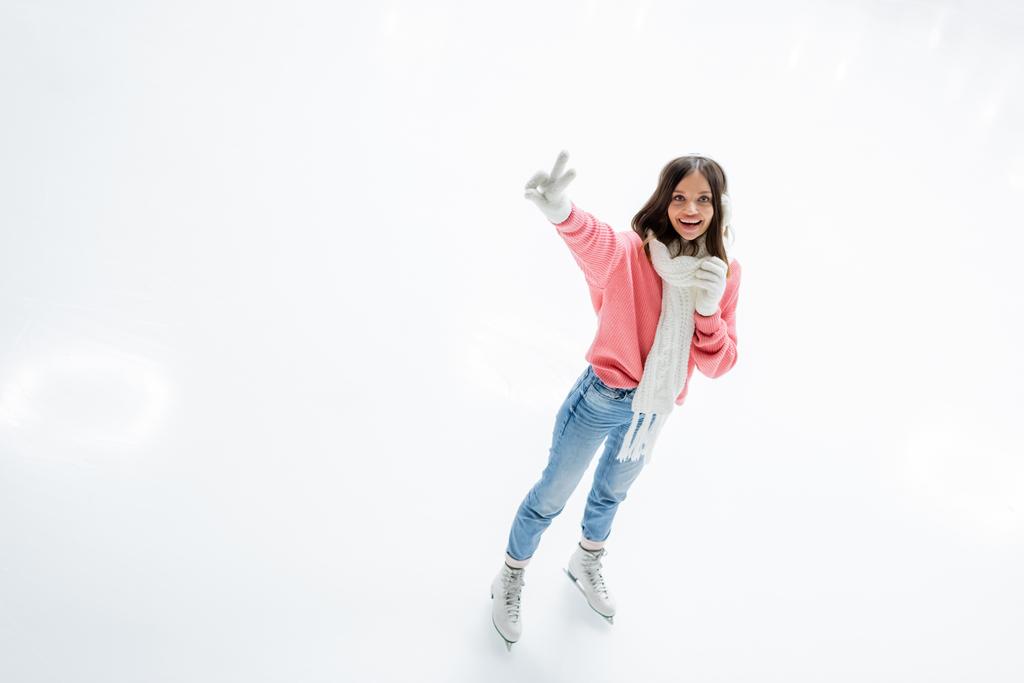 vista de ángulo alto de la joven feliz en orejeras y bufanda que muestra signo de paz mientras patina en pista de hielo - Foto, imagen
