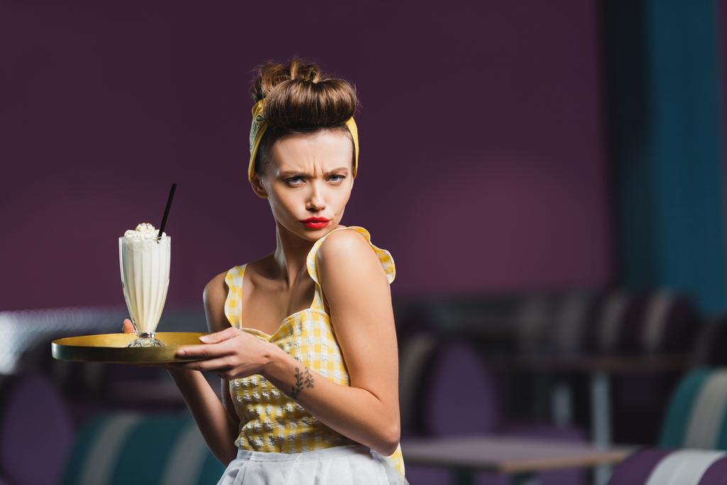displeased pin up waitress with tattoo holding tray and milkshake in cafe  - Photo, Image