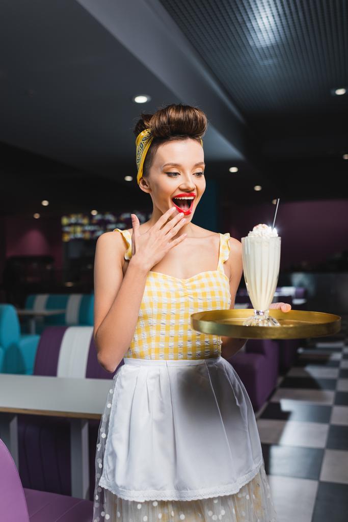zdumiony pin up kelnerka patrząc na tacę z milkshake w kawiarni  - Zdjęcie, obraz