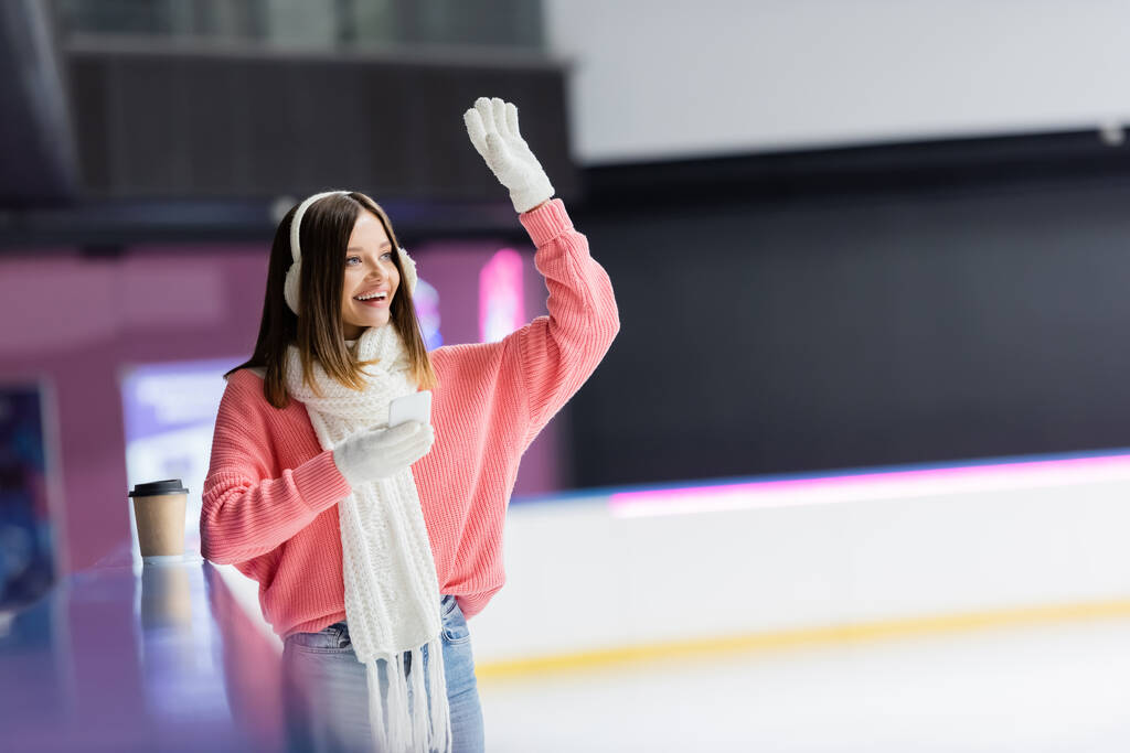 happy woman in ear muffs holding smartphone and waving hand on ice rink  - Photo, Image