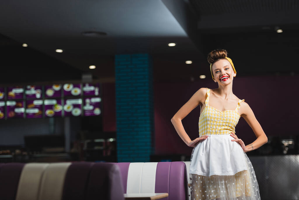 fröhliche Pin-up-Kellnerin steht mit den Händen auf den Hüften im Café  - Foto, Bild