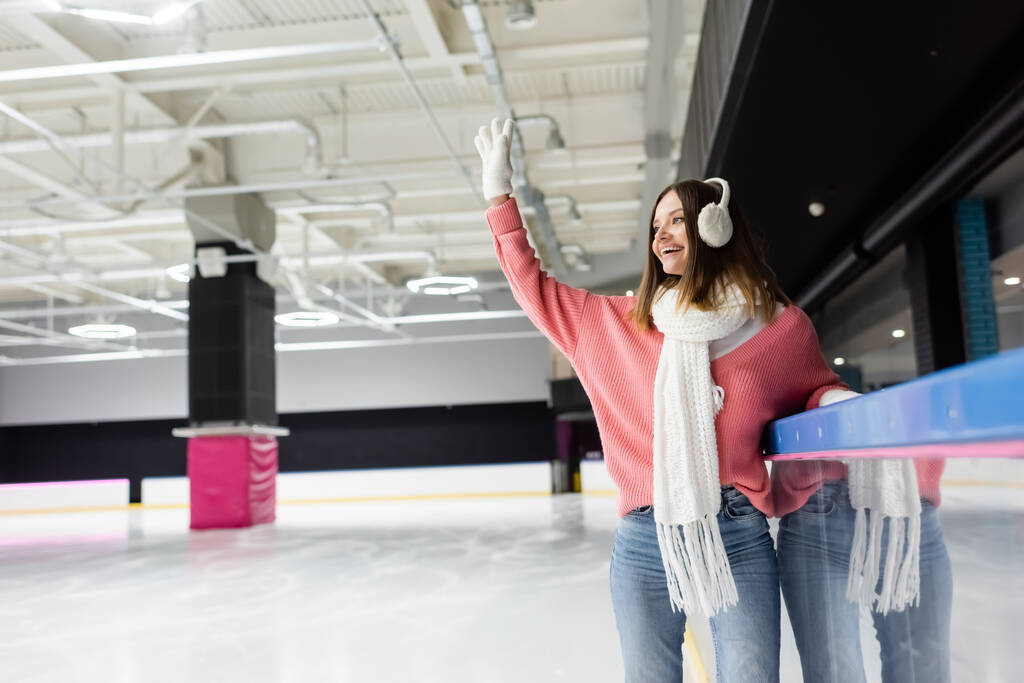 femme heureuse en gants blancs, cache-oreilles et pull rose agitant la main sur la patinoire - Photo, image