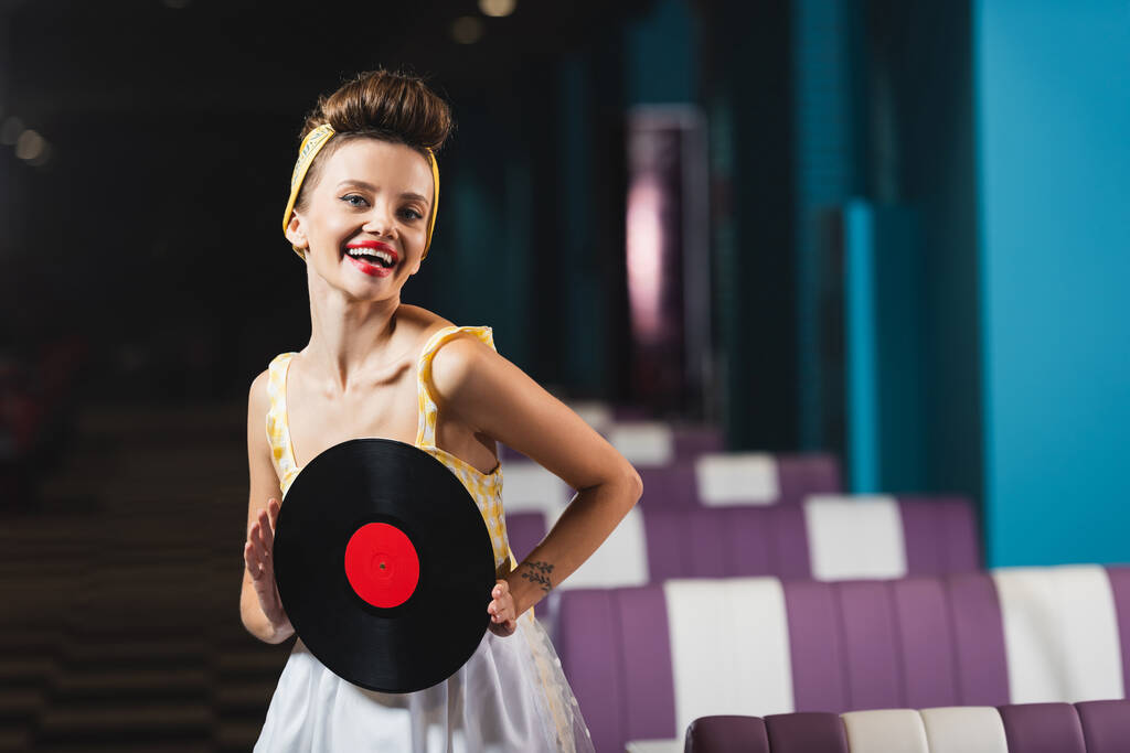 pin up žena s červenými rty drží retro vinylový disk a usmívá se v kavárně  - Fotografie, Obrázek