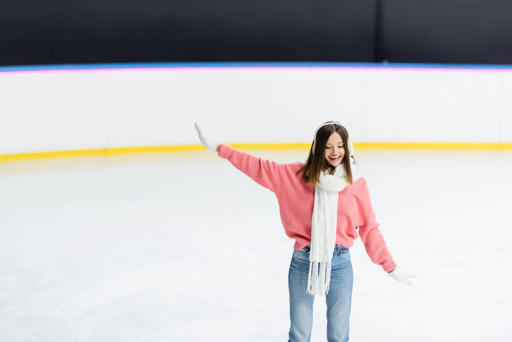 mujer sonriente en orejeras blancas, bufanda y suéter patinaje con la mano extendida en pista de hielo - Foto, Imagen