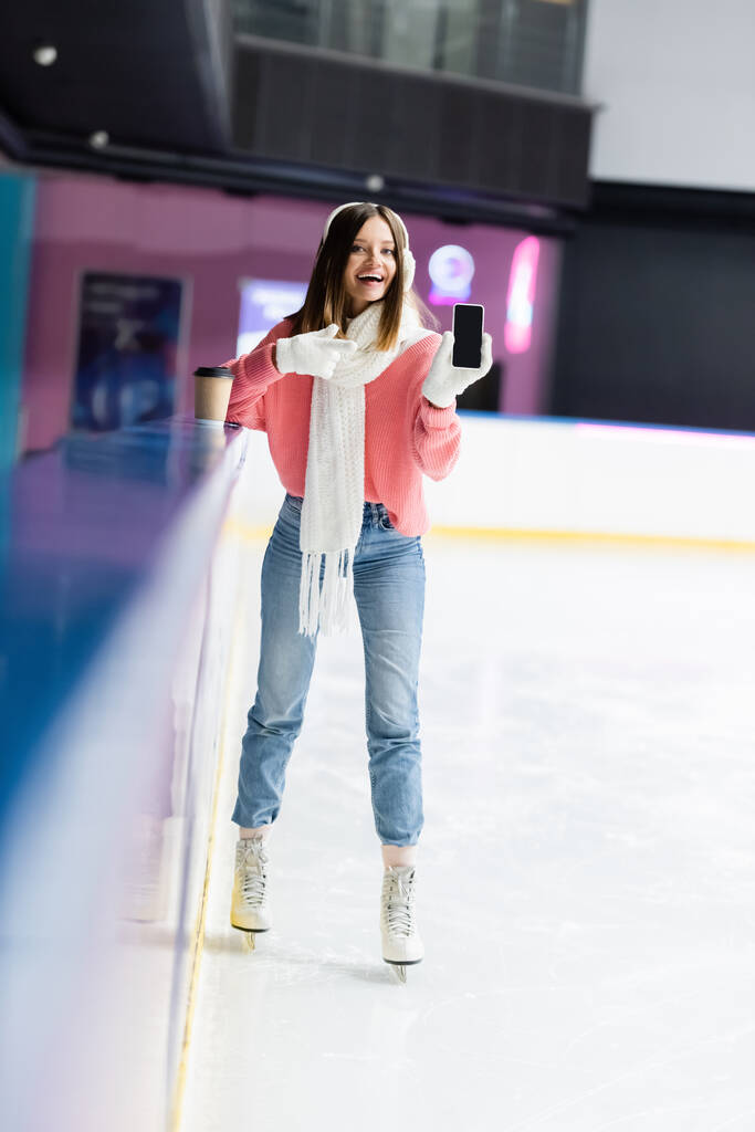 glückliche Frau in weißen Ohrenschützern und rosa Pullover zeigt mit dem Finger auf Handy mit leerem Bildschirm auf Eisbahn - Foto, Bild