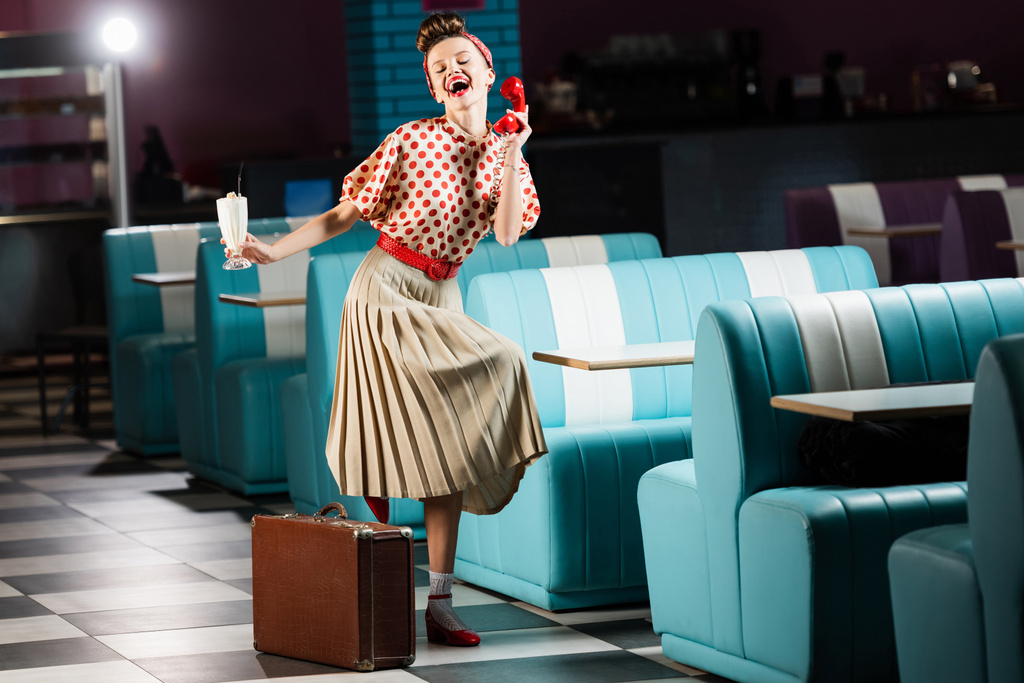amazed pin up woman holding milkshake cocktail and retro telephone near suitcase in cafe  - Photo, Image