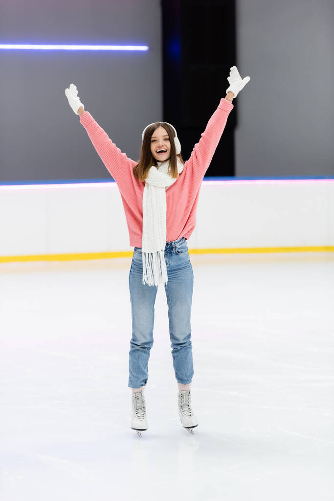 longitud completa de la mujer feliz en suéter de punto, orejeras y patinaje conjunto de invierno con las manos levantadas en la pista de hielo  - Foto, imagen