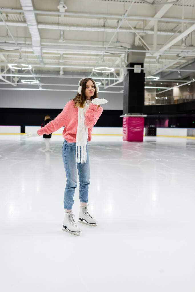 full length of happy woman in knitted sweater, ear muffs and winter outfit skating while sending air kiss on ice rink  - Photo, Image