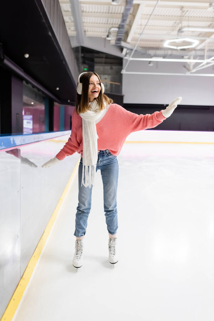 полная длина счастливой женщины в трикотажном свитере, наушниках и зимнем платье катание на коньках, размахивая рукой на катке  - Фото, изображение