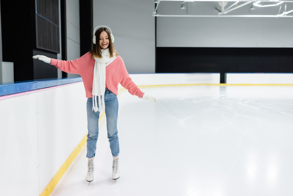 longitud completa de la mujer feliz en suéter de punto rosa, orejeras y patinaje conjunto de invierno en pista de hielo  - Foto, imagen