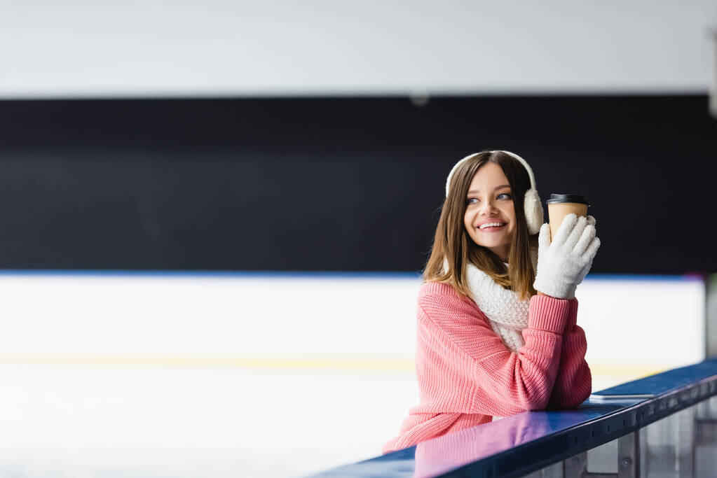 mujer feliz en suéter de punto, orejeras y traje de invierno sosteniendo taza de papel en pista de hielo - Foto, imagen