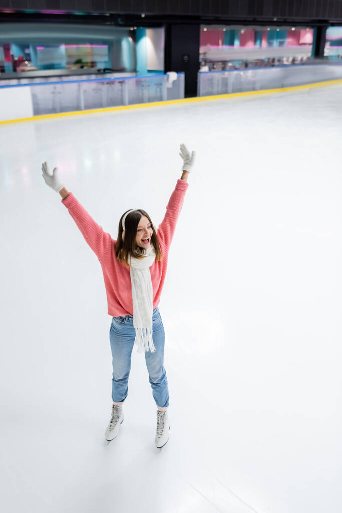 high angle view of amazed young woman in white ear muffs skating with raised hands on ice rink - Photo, Image