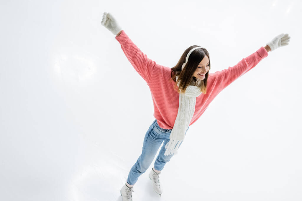 興奮した若い女性の耳のマフとジーンズスケートの高い角度ビューアイスリンクに手を伸ばし - 写真・画像