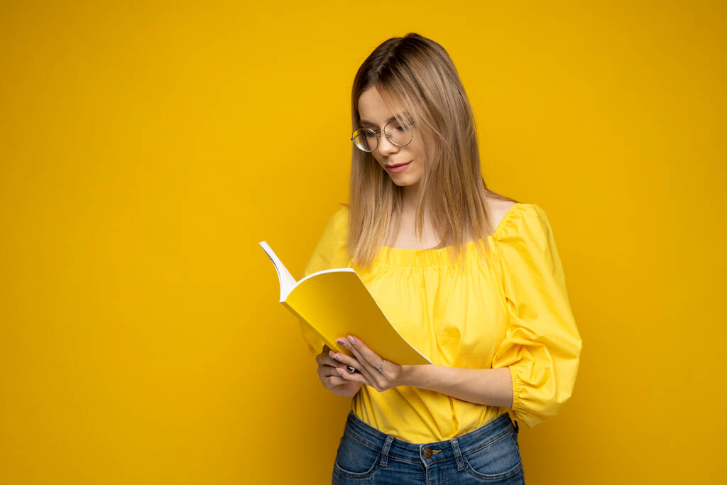 眼鏡をかけた若い十代の女性が黄色の背景に隔離された興味深い本を読んでいます。小説を読んでいる少女。勉強. - 写真・画像
