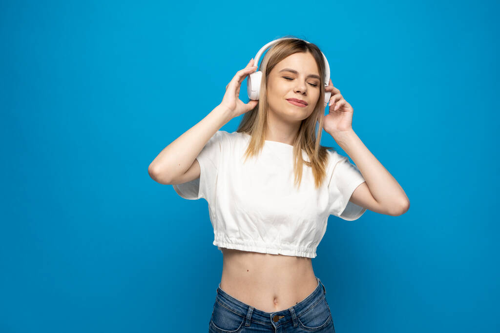 Красива молода жінка в навушниках слухає музичний синій фон. Молода красива жінка в яскравому вбранні насолоджується музикою
. - Фото, зображення