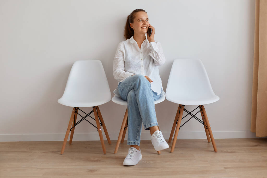 Ujęcie uśmiechniętej atrakcyjnej kobiety z kucykiem siedzącej na krześle przy lekkiej ścianie, kobiety w stylowych dżinsach i białej koszuli, rozmawiającej przez telefon komórkowy, odbywającej miłą rozmowę. - Zdjęcie, obraz