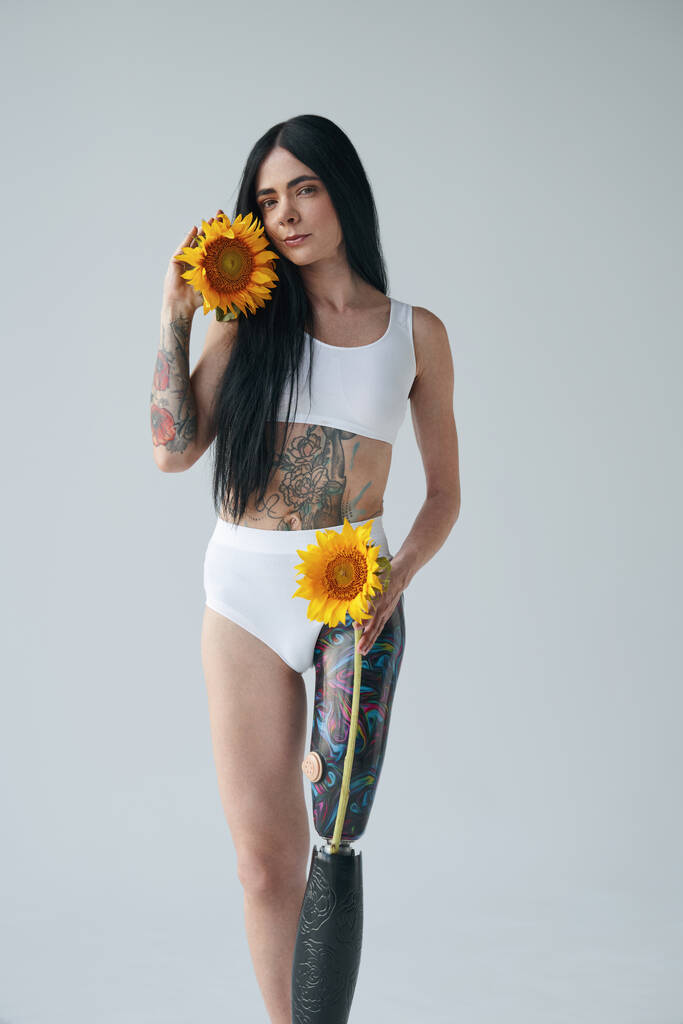 getatoeëerde vrouw met kunstbeen houden zonnebloemen en glimlachen zachtjes - Foto, afbeelding
