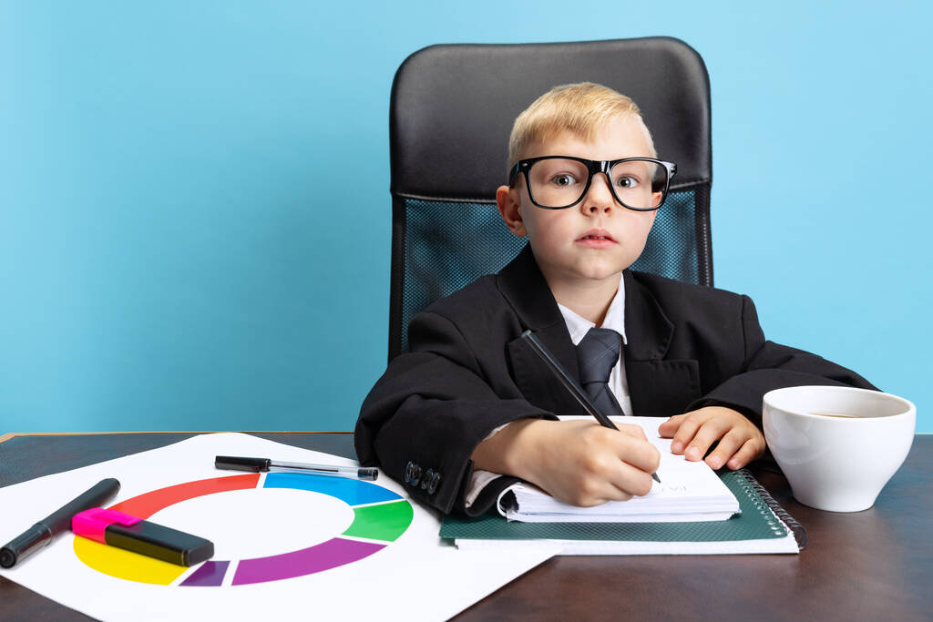 Portrait de petit garçon, enfant homme d'affaires en costume de veste énorme assis à la table de bureau isolé sur fond bleu studio. - Photo, image