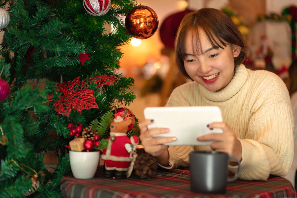 Schöne Frau im Pullover lächelnd und auf einem Stuhl sitzend, um mit einem Freund auf einem Tablet in der Nähe des Weihnachtsbaums zu plaudern, dekoriert mit glitzernden Kugeln und Dekorationen für die Feier von Weihnachten oder Neujahr - Foto, Bild