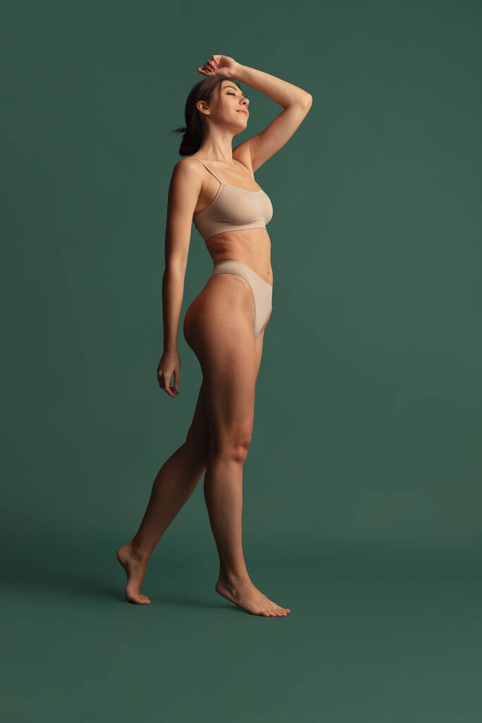 Porträt einer jungen schönen schlanken Frau in Unterwäsche in Nudefarben, die isoliert vor dunkelgrünem Studiohintergrund posiert. Natürliches Schönheitskonzept. - Foto, Bild