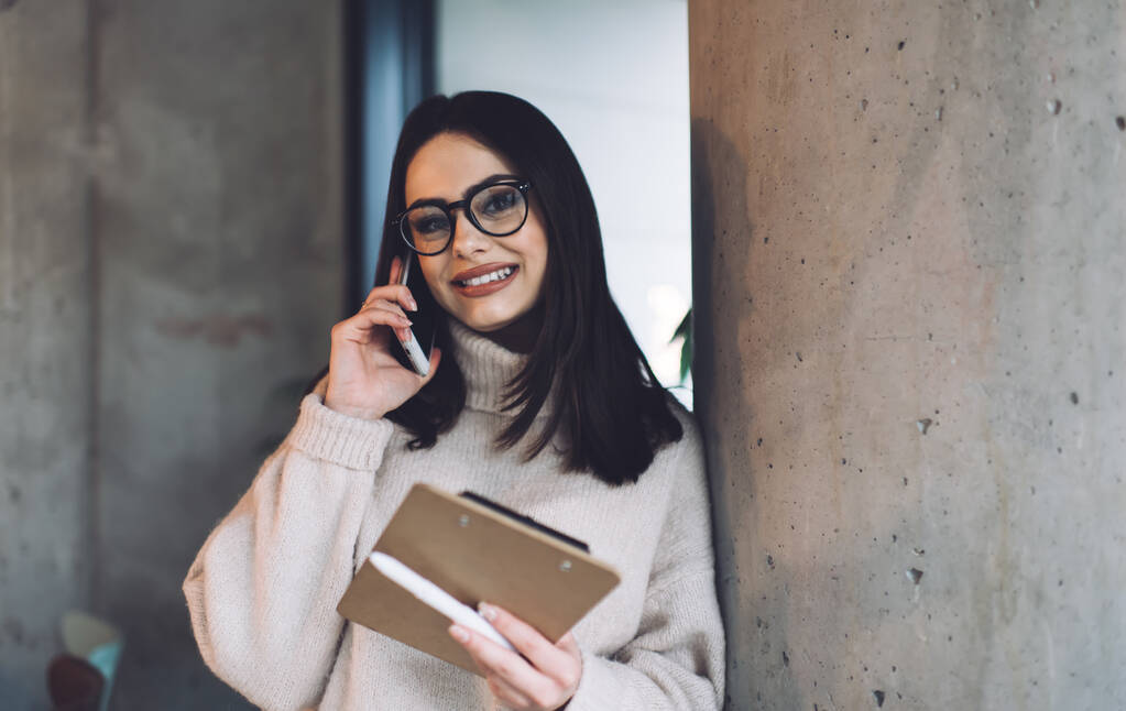 Szczęśliwa kobieta w luźnych ubraniach i okularach uśmiechnięta i patrząca w kamerę podczas rozmowy na smartfonie stojąc w nowoczesnym biurze - Zdjęcie, obraz