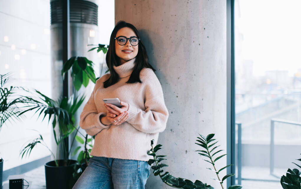 Веселая женщина в повседневном свитере и очках, стоящая в современном офисе и отворачивающаяся от сообщений на смартфоне, улыбаясь - Фото, изображение