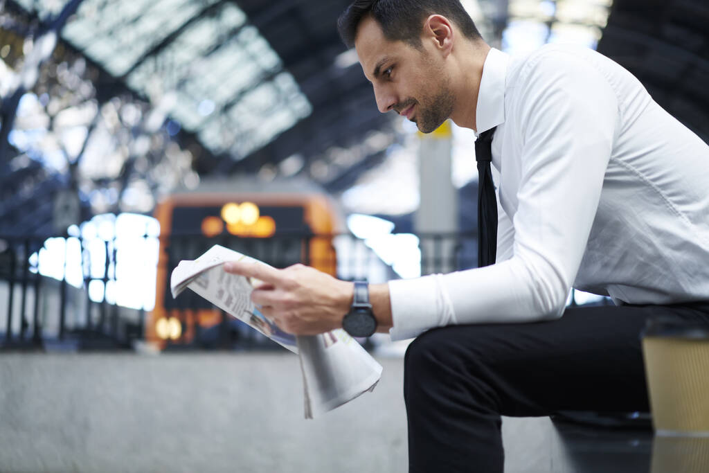Вид сбоку на уверенного предпринимателя в формальном костюме, сидящего на скамейке и наслаждающегося чтением газет в ожидании поезда - Фото, изображение