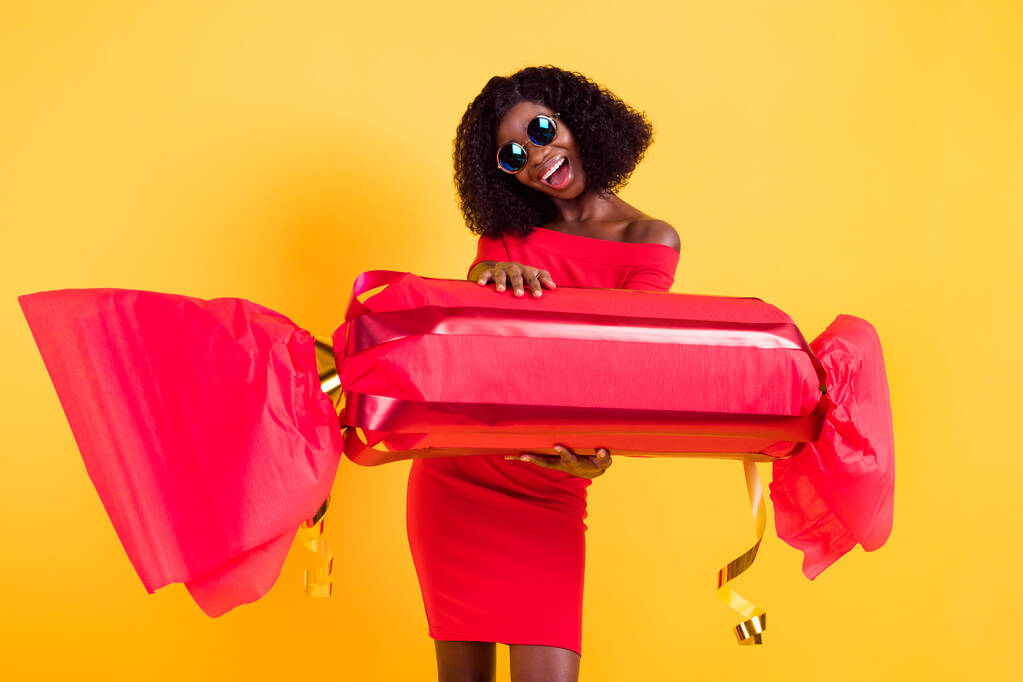 Фото молодой очаровательной возбужденной позитивной улыбающейся афро-девушки в солнечных очках провести огромные конфеты изолированы на желтом фоне цвета - Фото, изображение