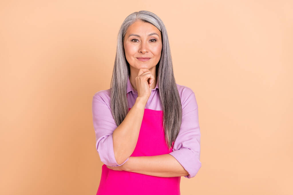 Фото розумного сірого волосся старша леді думає носити фіолетову сорочку рожевий фартух ізольовані на бежевому кольоровому фоні
 - Фото, зображення