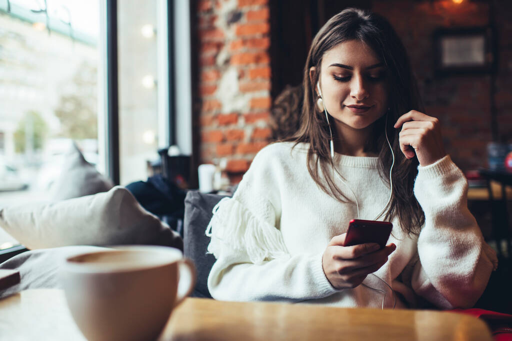 Jovem fêmea sorridente em roupas casuais sentada à mesa perto da janela conversando no celular e ouvindo música no café - Foto, Imagem