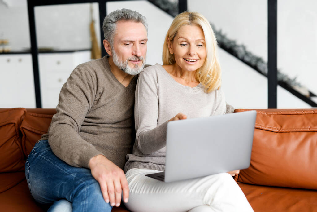 Joyeux couple d'âge moyen avec un ordinateur portable assis sur le canapé en cuir confortable - Photo, image
