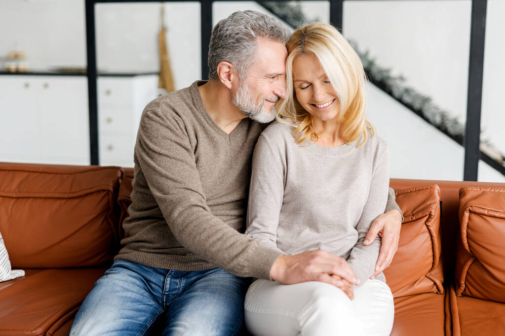 Lovely middle-aged couple in love sitting in embrace on the comfortable sofa - Photo, Image