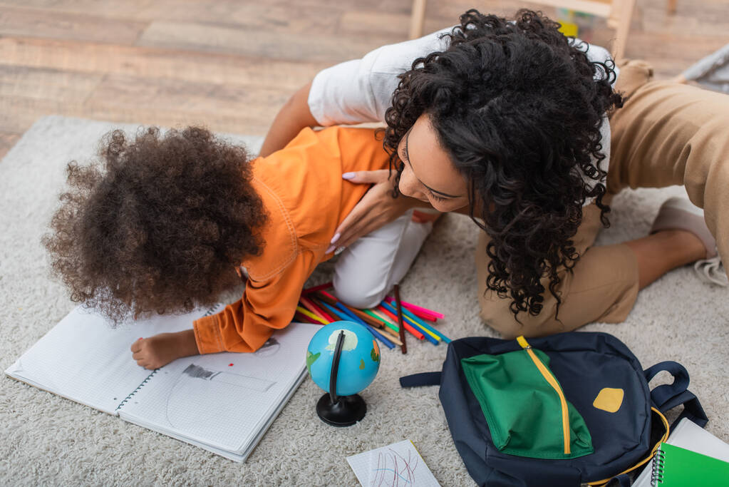 Afro-Amerikaanse moeder knuffelen kind tekening op notebook in de buurt van globe en rugzak  - Foto, afbeelding