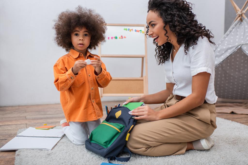 Feliz mãe afro-americana olhando para a filha perto de cadernos e mochila na sala de estar  - Foto, Imagem