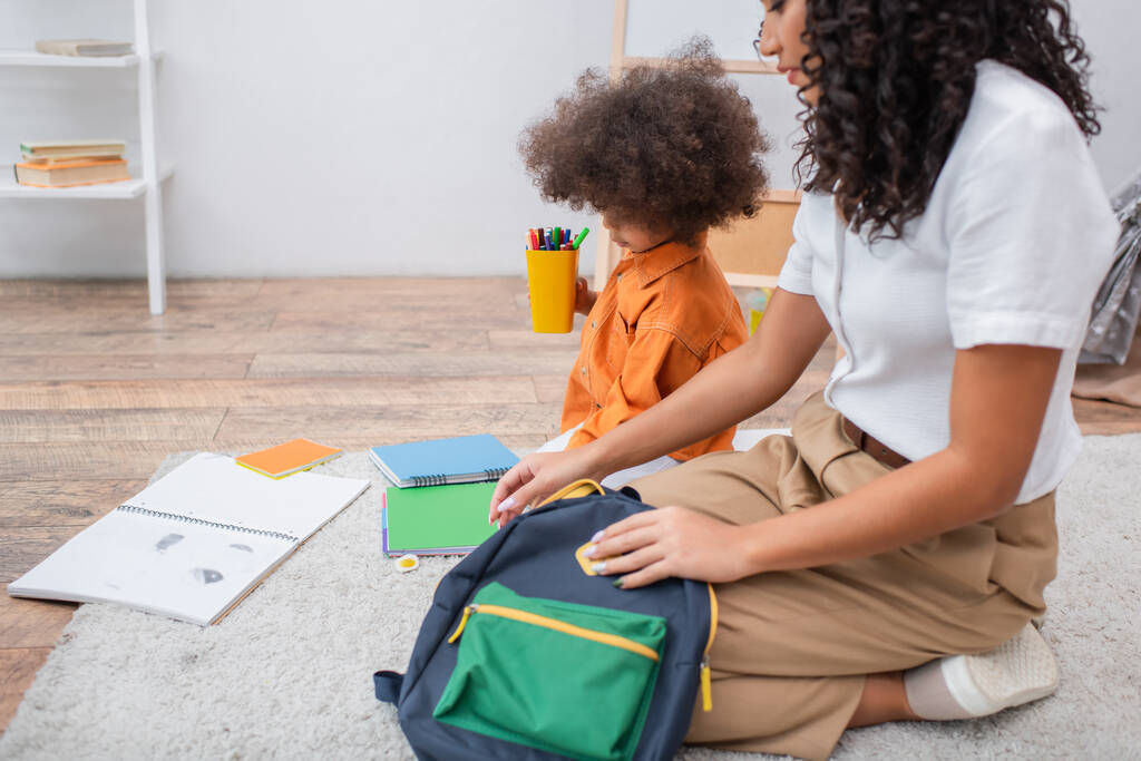 Африканський американець тримає кольорові олівці біля зошитів і мати з рюкзаком вдома.  - Фото, зображення