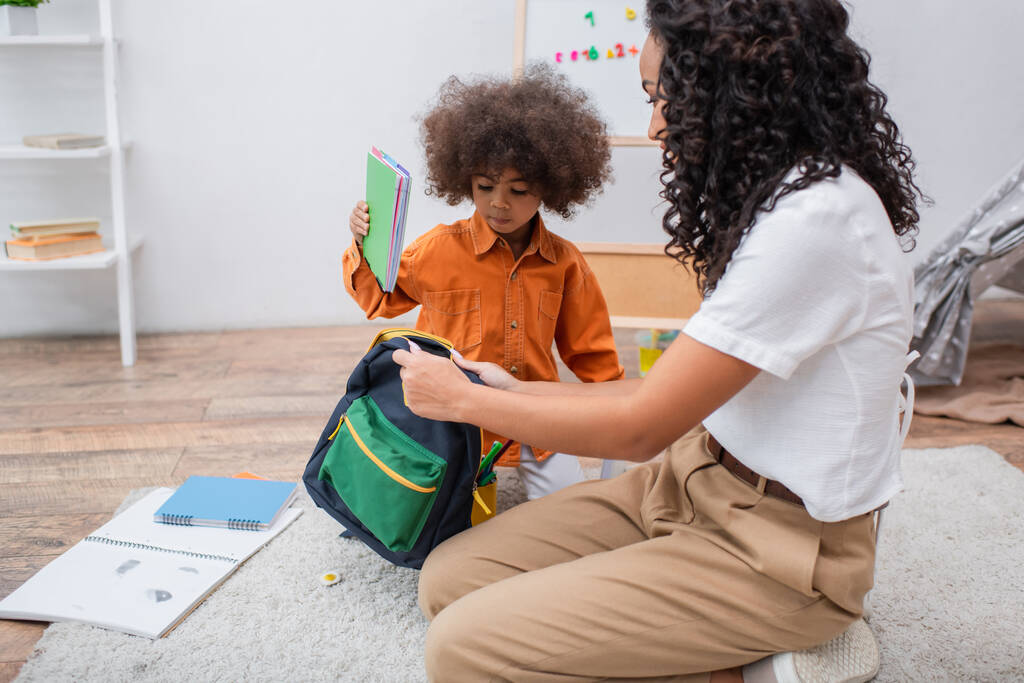 Afrikanisches Kind hält Notizbuch neben Mutter mit Rucksack im Wohnzimmer  - Foto, Bild