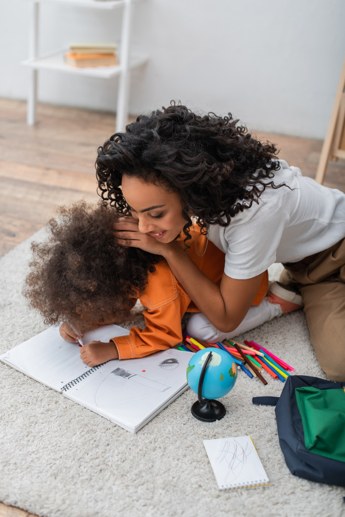 Young african american mom sitting near daughter drawing on notebook and globe at home  - 写真・画像